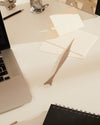 Silver fish-shaped letter opener by Alessi, laying diagonally on a white desk beside white envelopes and a black notebook.