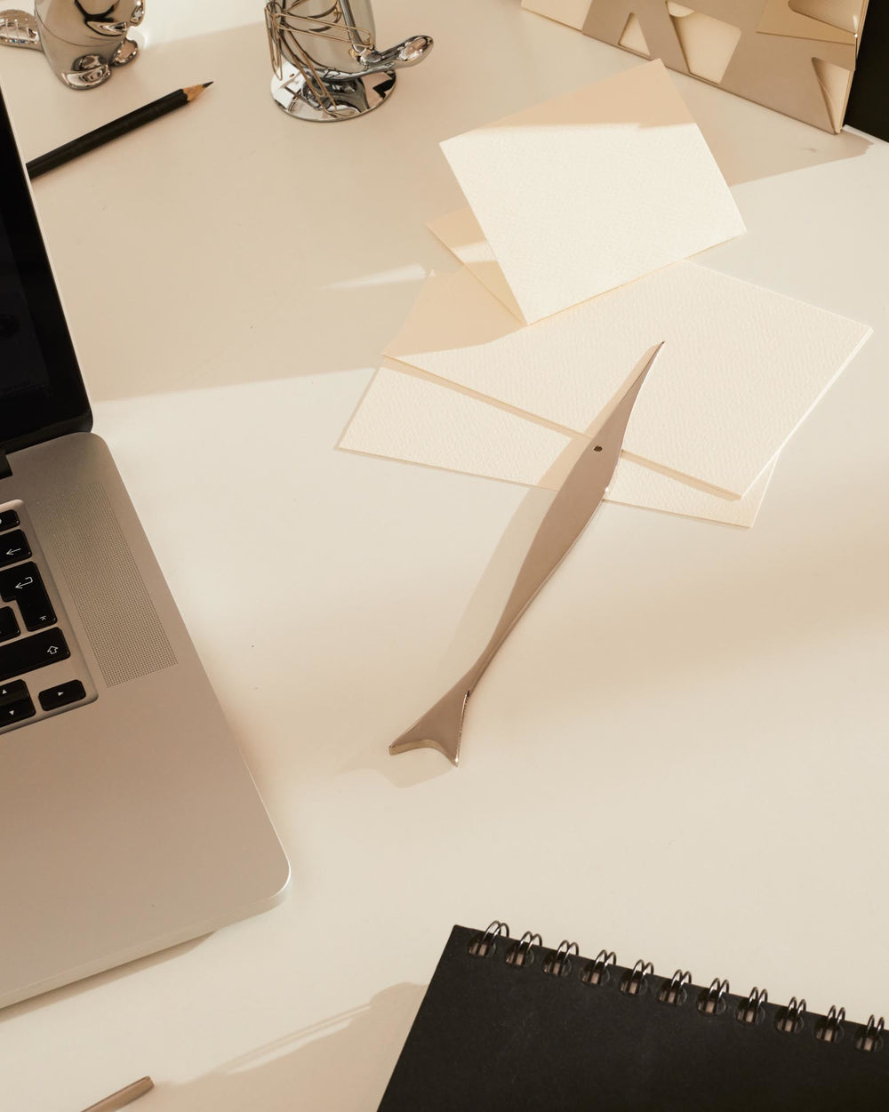 Silver fish-shaped letter opener by Alessi, laying diagonally on a white desk beside white envelopes and a black notebook.