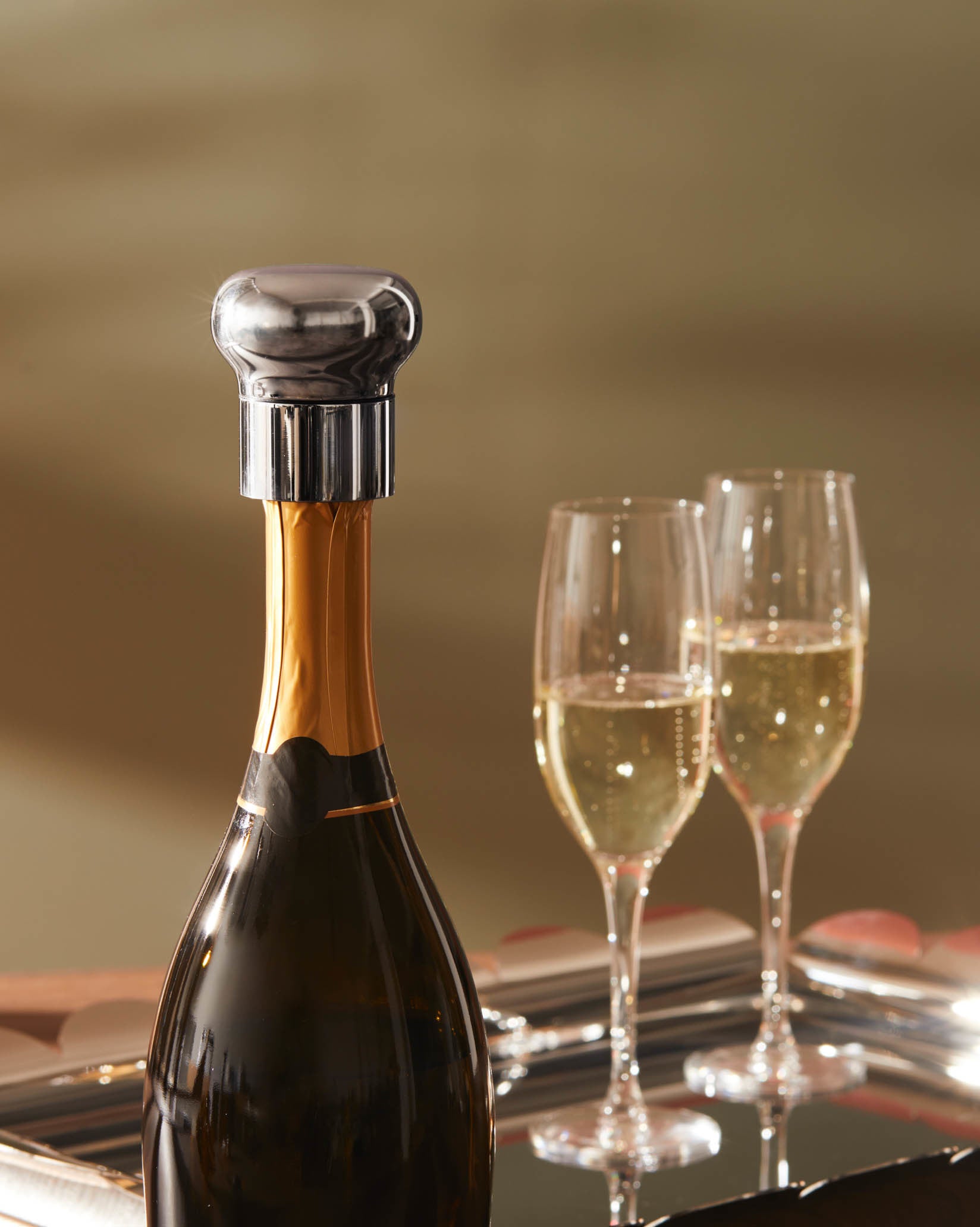Silver polished metal bottle stopper by Alessi, with a bulbous top and ribbed base, on a champagne bottle with glasses.