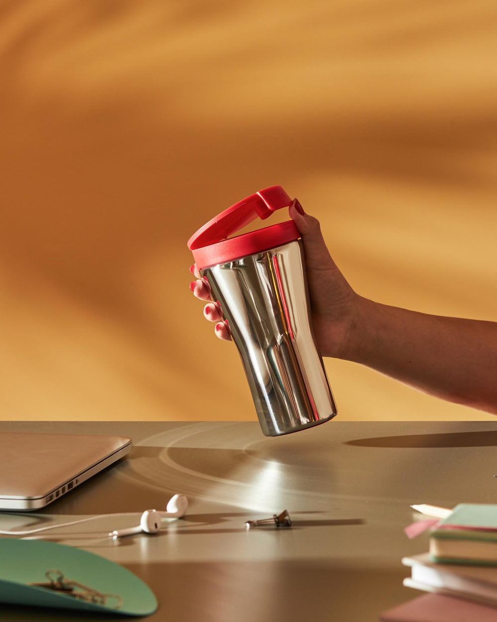 Silver stainless steel travel mug by Alessi with a red lid, held by a hand with red nail polish on a desk.