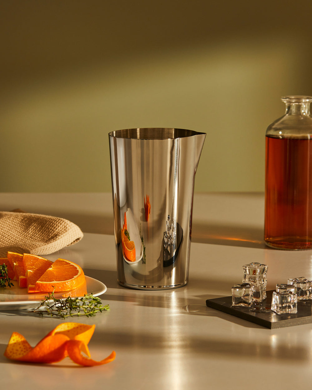 Sleek stainless steel cocktail mixing jug by Alessi, reflecting orange slices, on a countertop with ice and spirits.