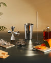 Silver metallic cocktail tending box by Alessi, featuring a jigger, shaker, and muddler on a dark surface with ice and oranges.