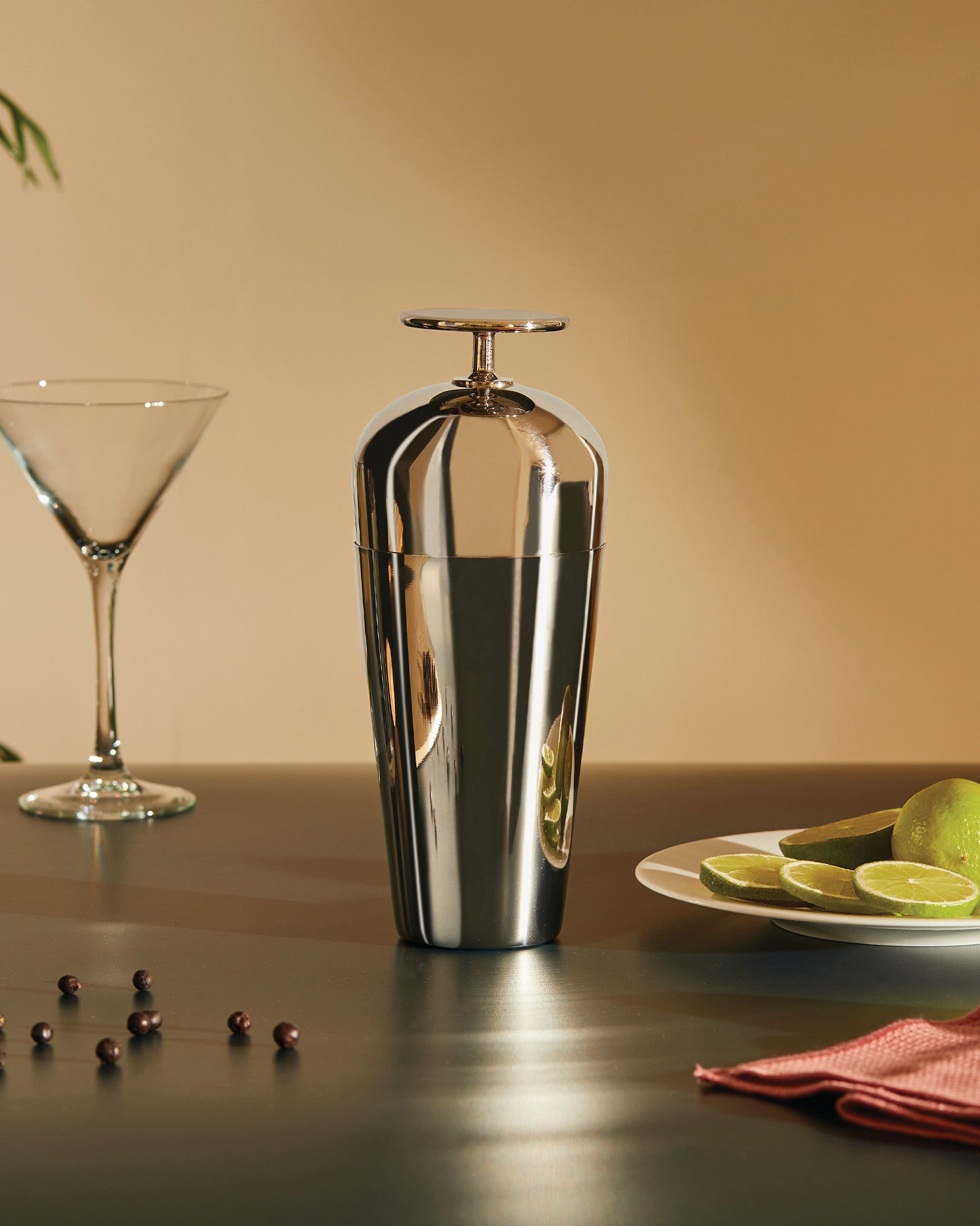 Shiny metallic cocktail shaker by Alessi, centered on a dark surface, beside a martini glass and limes.