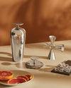 Polished silver cocktail shaker set by Alessi, featuring a sleek shaker, unique cross jigger, and round strainer.