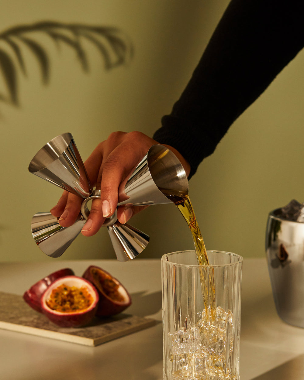 The tending box by Alessi, featuring a stainless steel jigger pouring golden liquid into a fluted glass with ice and passion fruit.