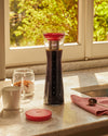 Tall glass cold brew coffee pitcher by Alessi with a red filter and lid.