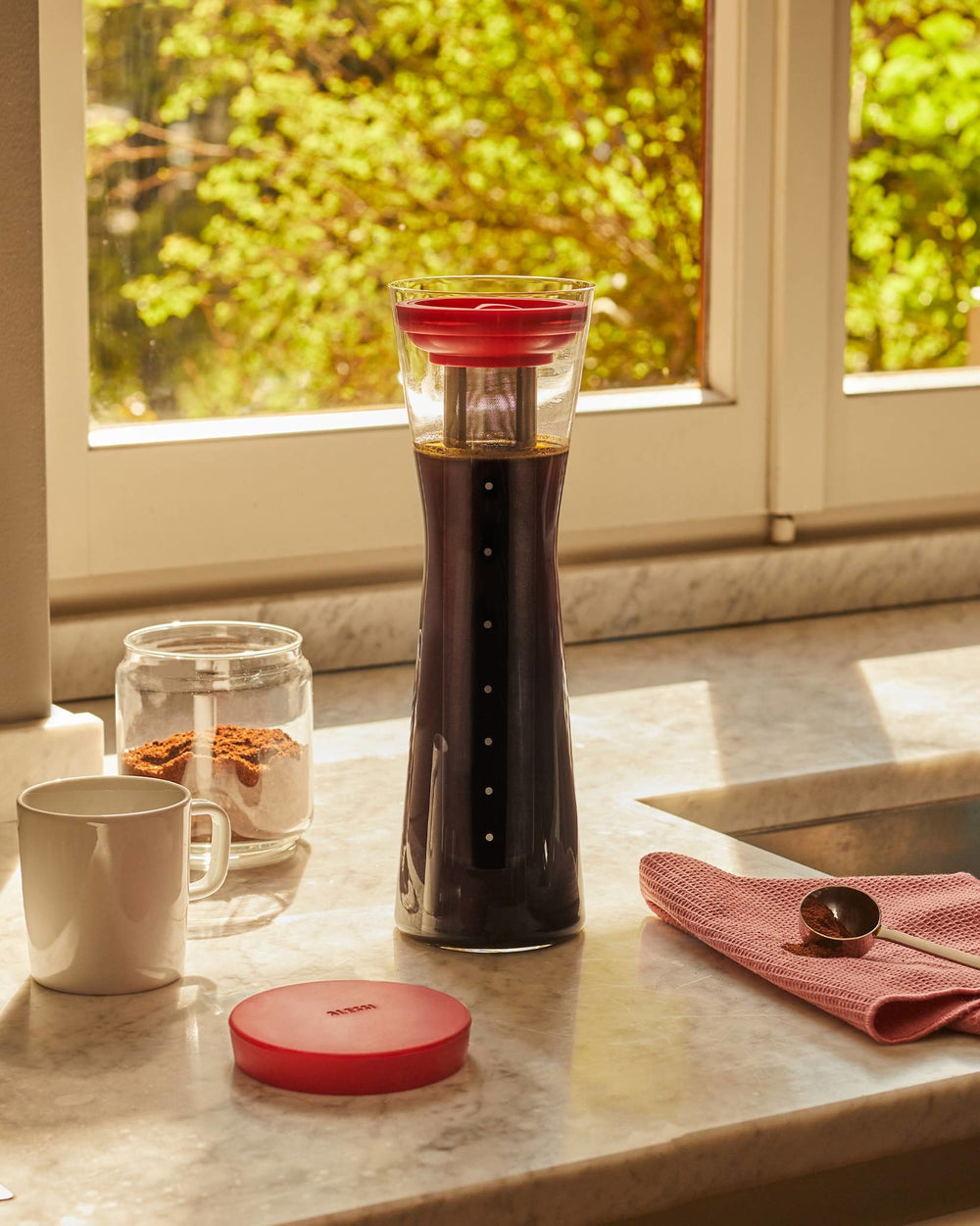 Tall glass cold brew coffee pitcher by Alessi with a red filter and lid.