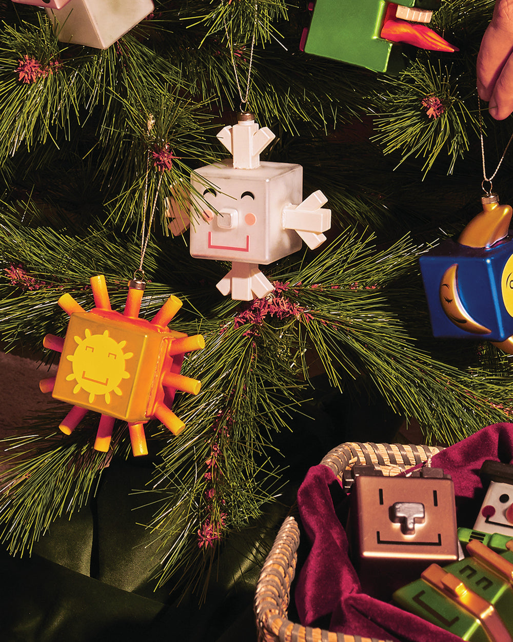 Orange Sunflake ornament by Alessi, a cuboid sun with a smiling face, hanging on a Christmas tree.