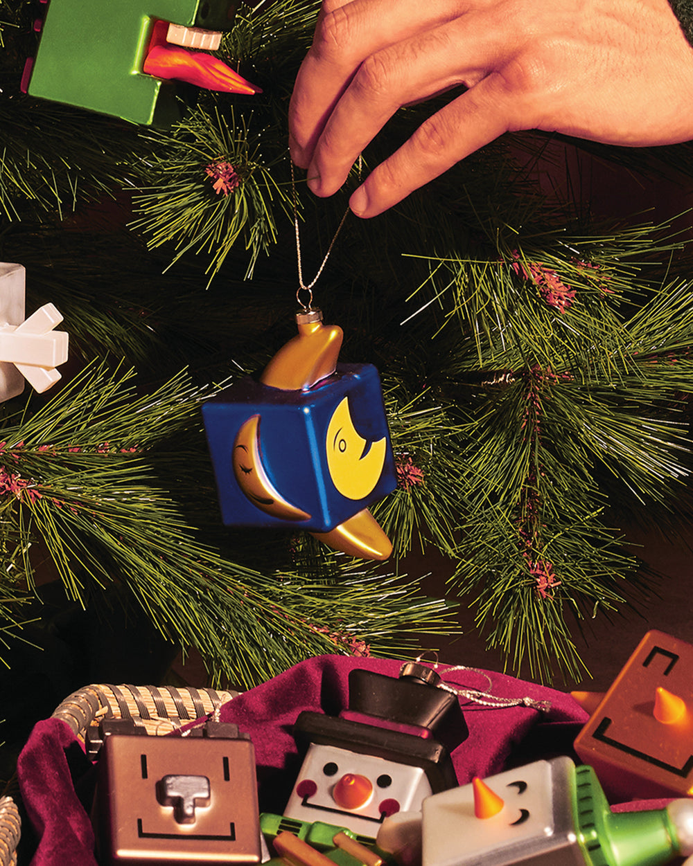 Blue and gold cube-shaped Mooncube ornament by Alessi, featuring crescent and full moon designs, being hung on a green Christmas tree.