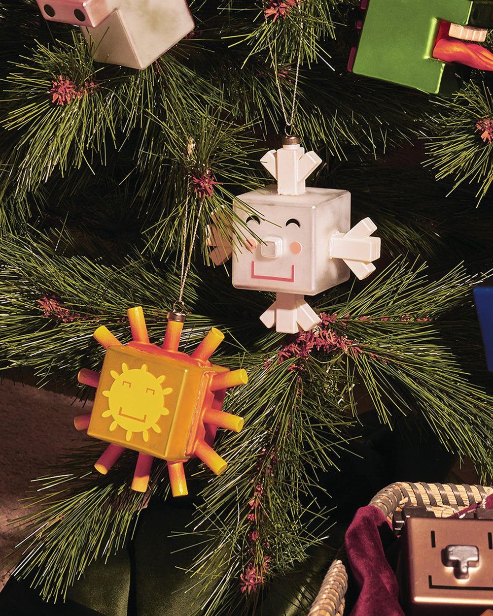 White cube-shaped Snowray ornament by Alessi, with a smiling face, hanging on a Christmas tree next to an orange sun ornament.