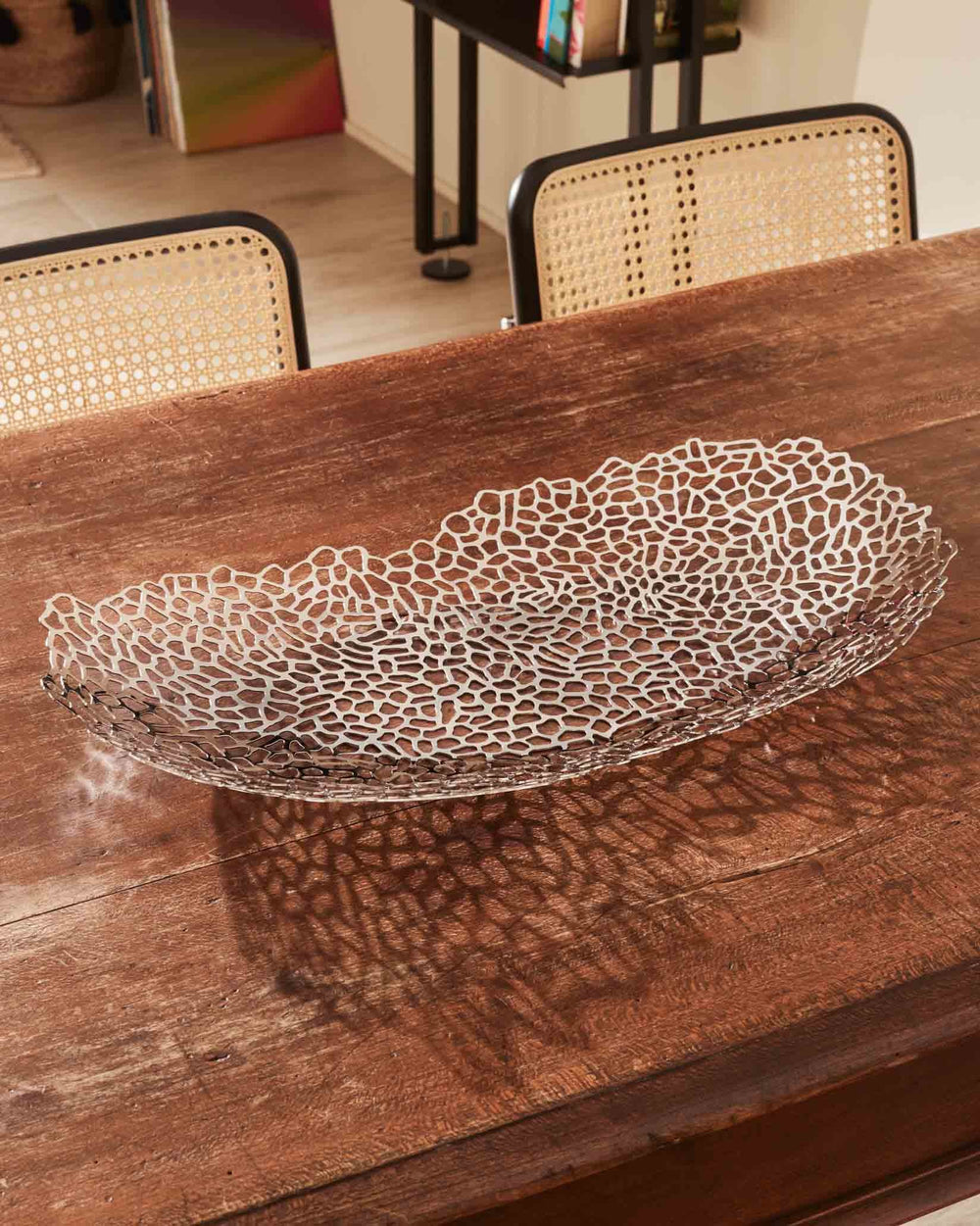 Silver oval centrepiece by Alessi featuring an intricate organic openwork design, casting shadows on a rustic wooden table.