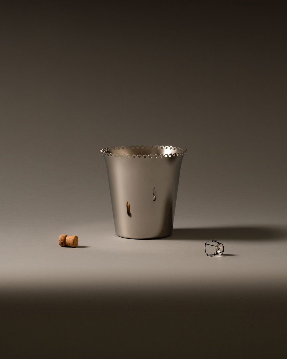 Silver wine cooler by Alessi with a perforated top edge, displayed with a cork and wire cage on a grey surface.