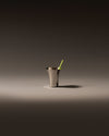 Brushed metal glass by Alessi with a flared rim and a green straw, on a grey surface.