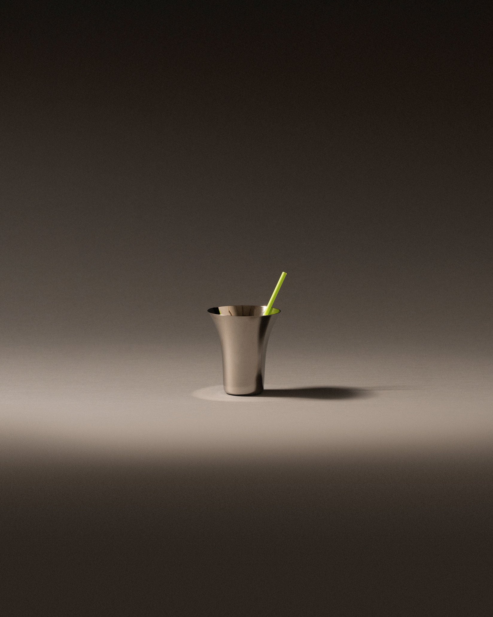 Brushed metal glass by Alessi with a flared rim and a green straw, on a grey surface.