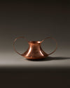 Copper Conca vase by Alessi with wide, circular handles on a dark background.