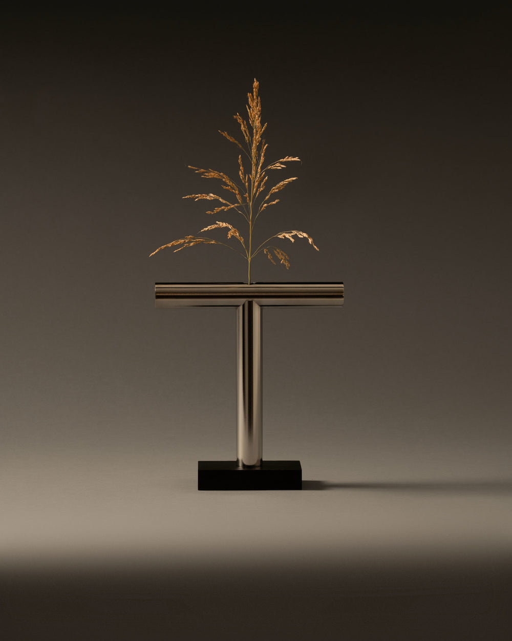 T-shaped metallic vase by Alessi on a black base, holding a dried golden plant.