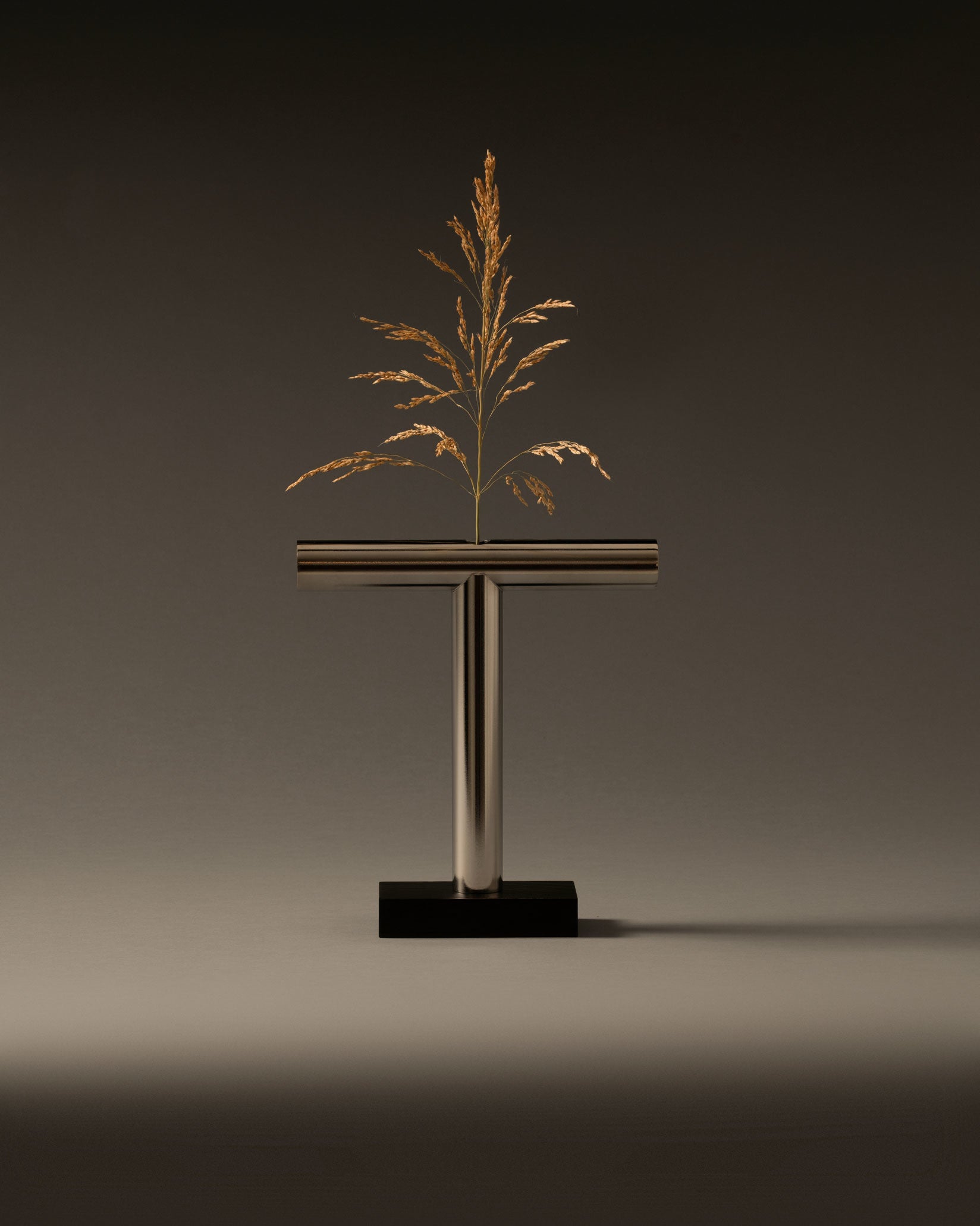 T-shaped metallic vase by Alessi on a black base, holding a dried golden plant.
