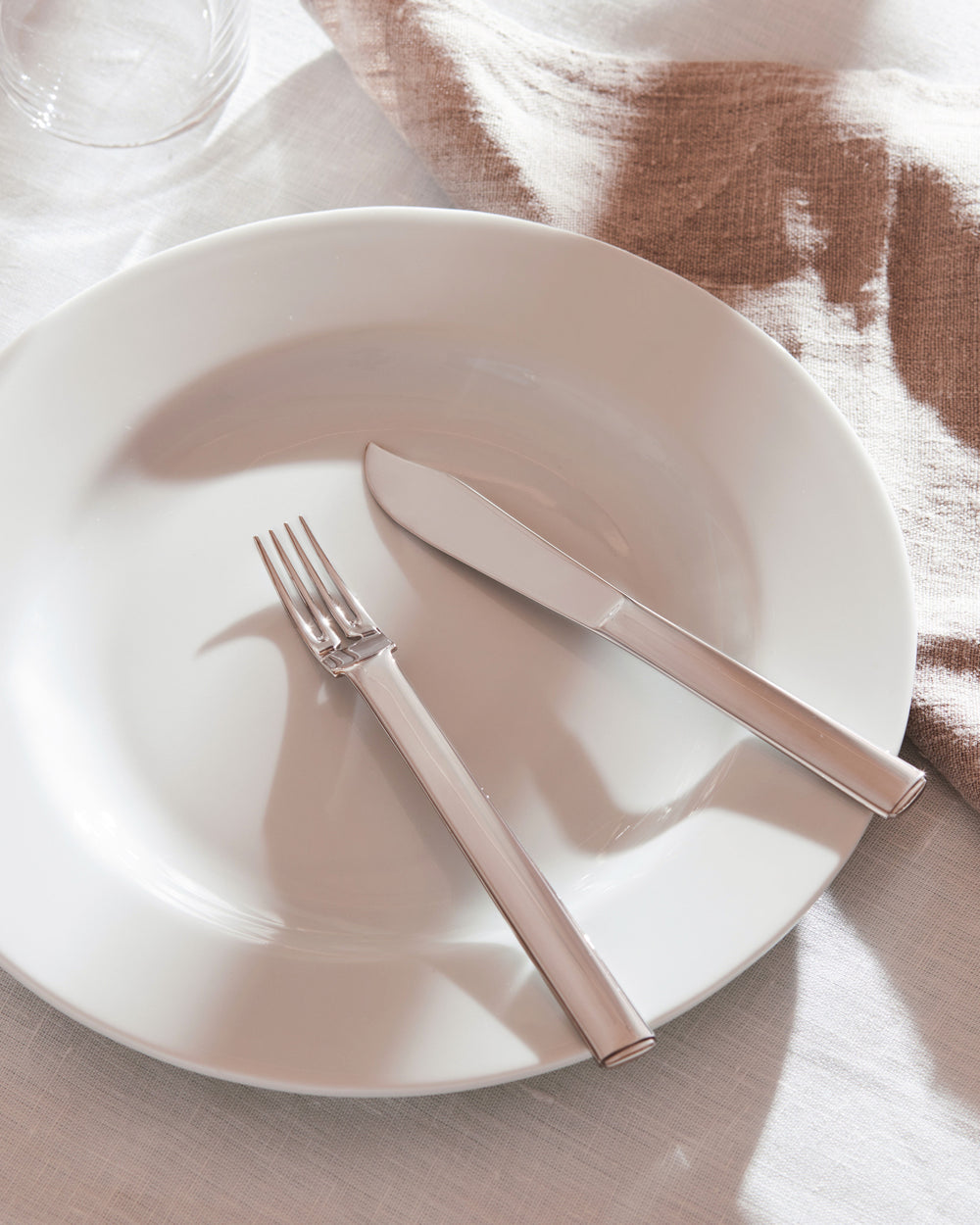 Shiny silver cutlery set by Alessi, featuring a fork and knife placed on a white plate with a textured brown napkin.