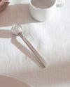 Silver round-bowled spoon by Alessi, polished metal, lying on a white linen tablecloth beside a white mug.