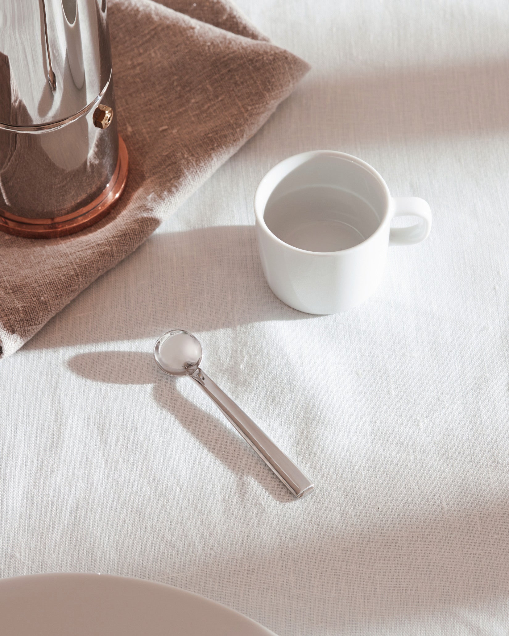 Silver moka pot by Alessi, with a copper base, on a linen napkin next to a white cup and spoon.