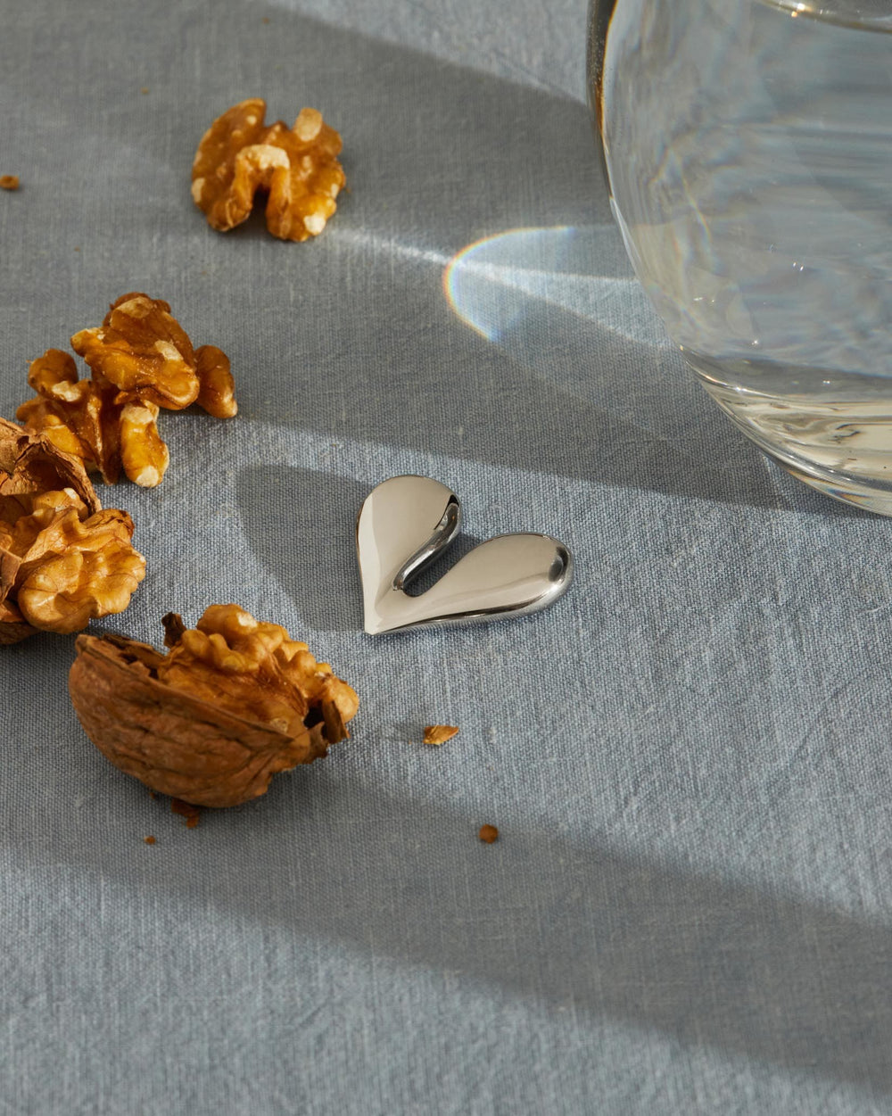 Sleek silver heart-shaped NUT SPLITTER by Alessi, lying on a blue fabric surrounded by walnuts.