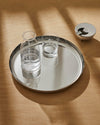 A round silver serving tray by Alessi holding a glass carafe and drinking glass, with a metal ashtray on a wooden table.
