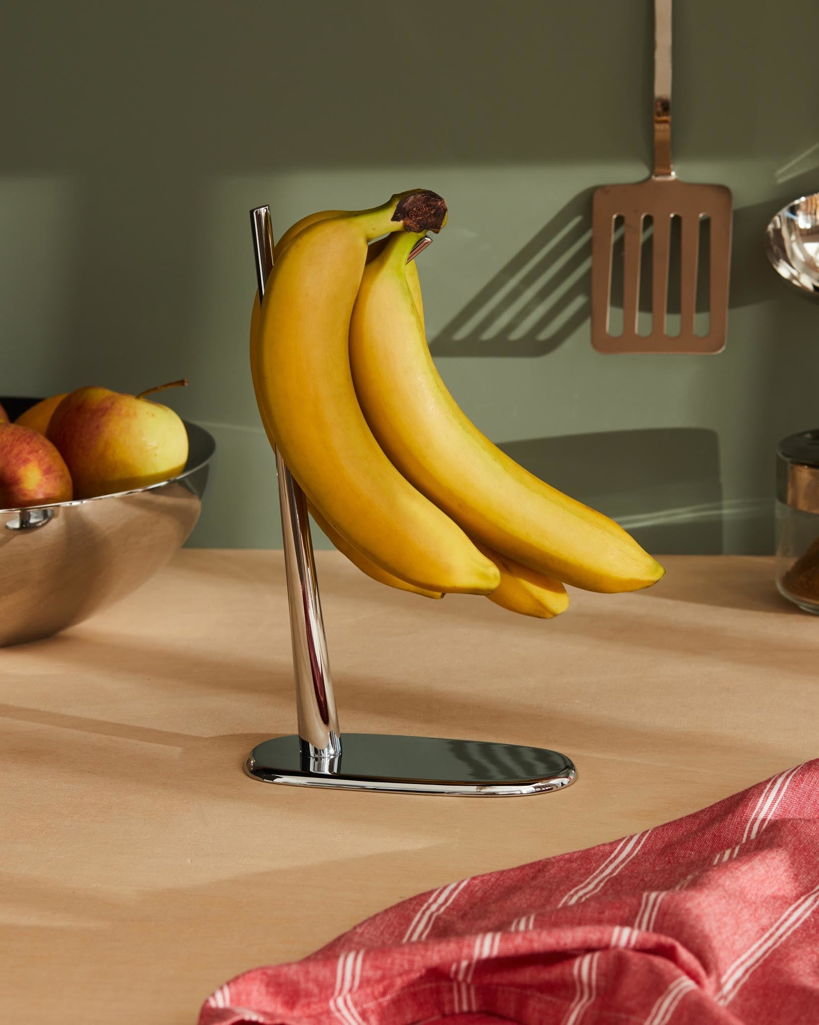 Polished stainless steel fruit bowl by Alessi, with apples, next to bananas on a stand and a red striped towel on a counter.