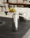 Sleek stainless steel coffee grinder by Alessi with a black handle, on a dark countertop next to a jar of coffee beans.