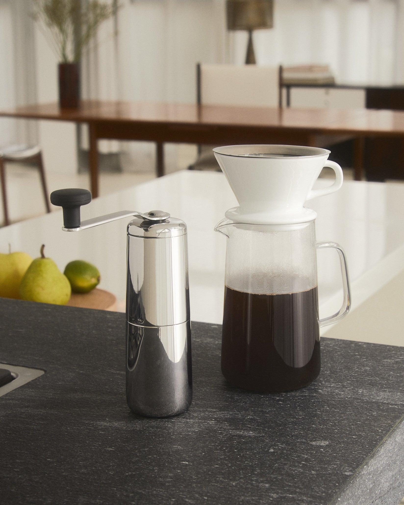 Chrome manual coffee grinder by Alessi with black handle, shown next to a glass pour-over coffee carafe and fruit.
