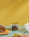 Clear glass storage jar by Alessi with a shiny gold lid and hazelnuts, on a green table with bread, a white bowl, and flowers.