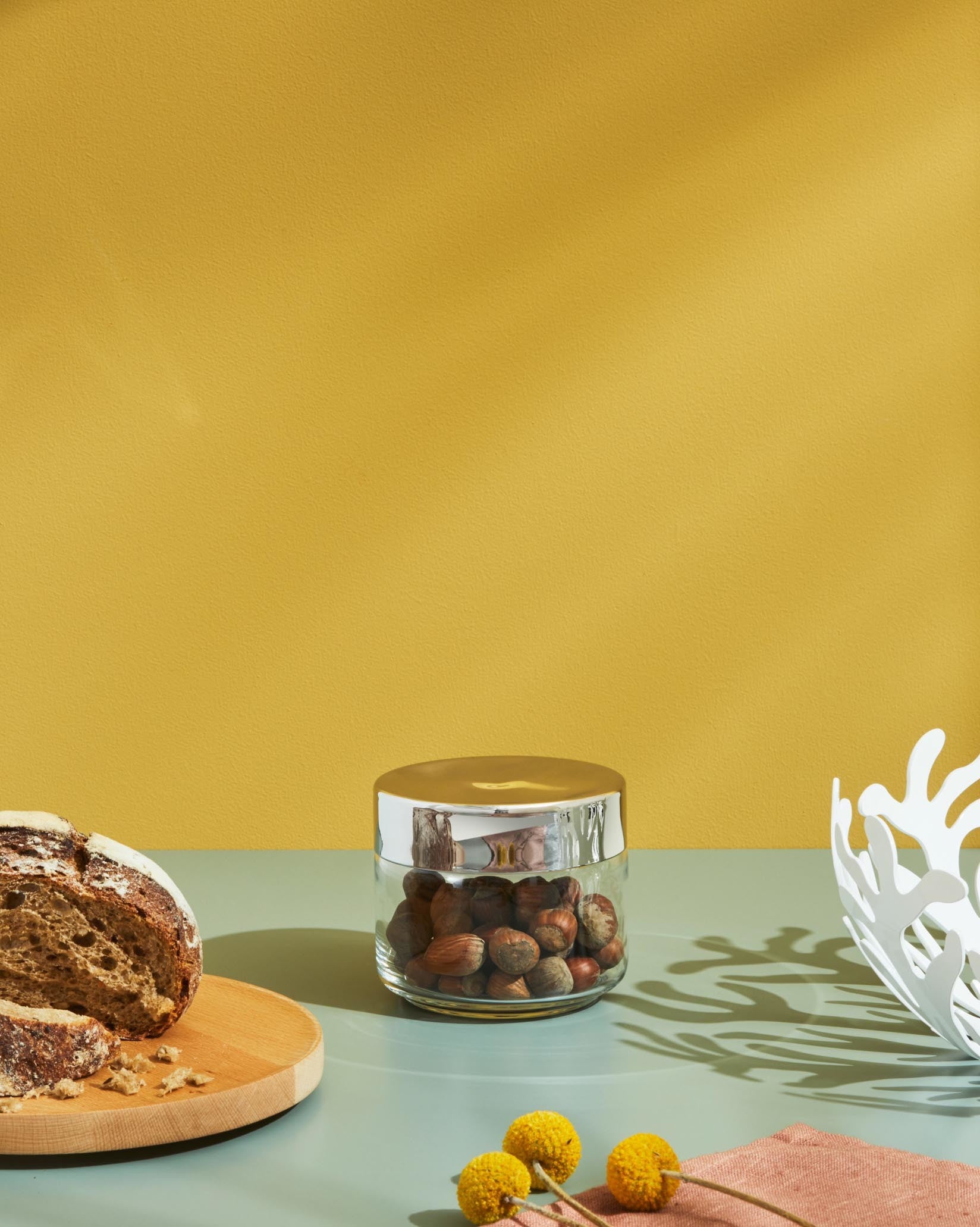 Clear glass storage jar by Alessi with a shiny gold lid and hazelnuts, on a green table with bread, a white bowl, and flowers.