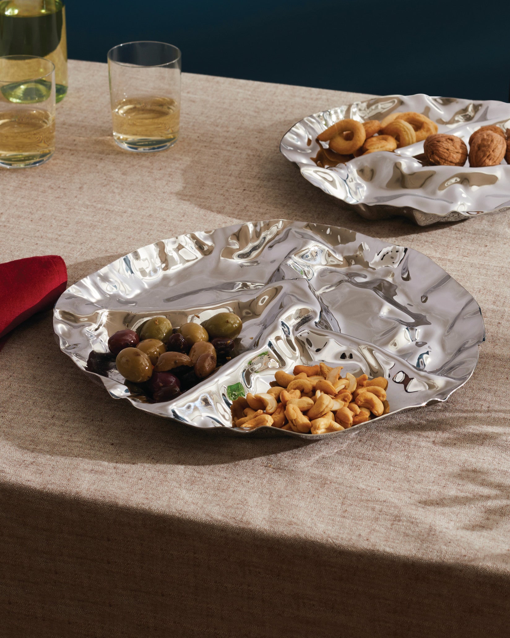 Silver-toned, crinkled serving plates by Alessi, one segmented with olives and cashews on a beige tablecloth.
