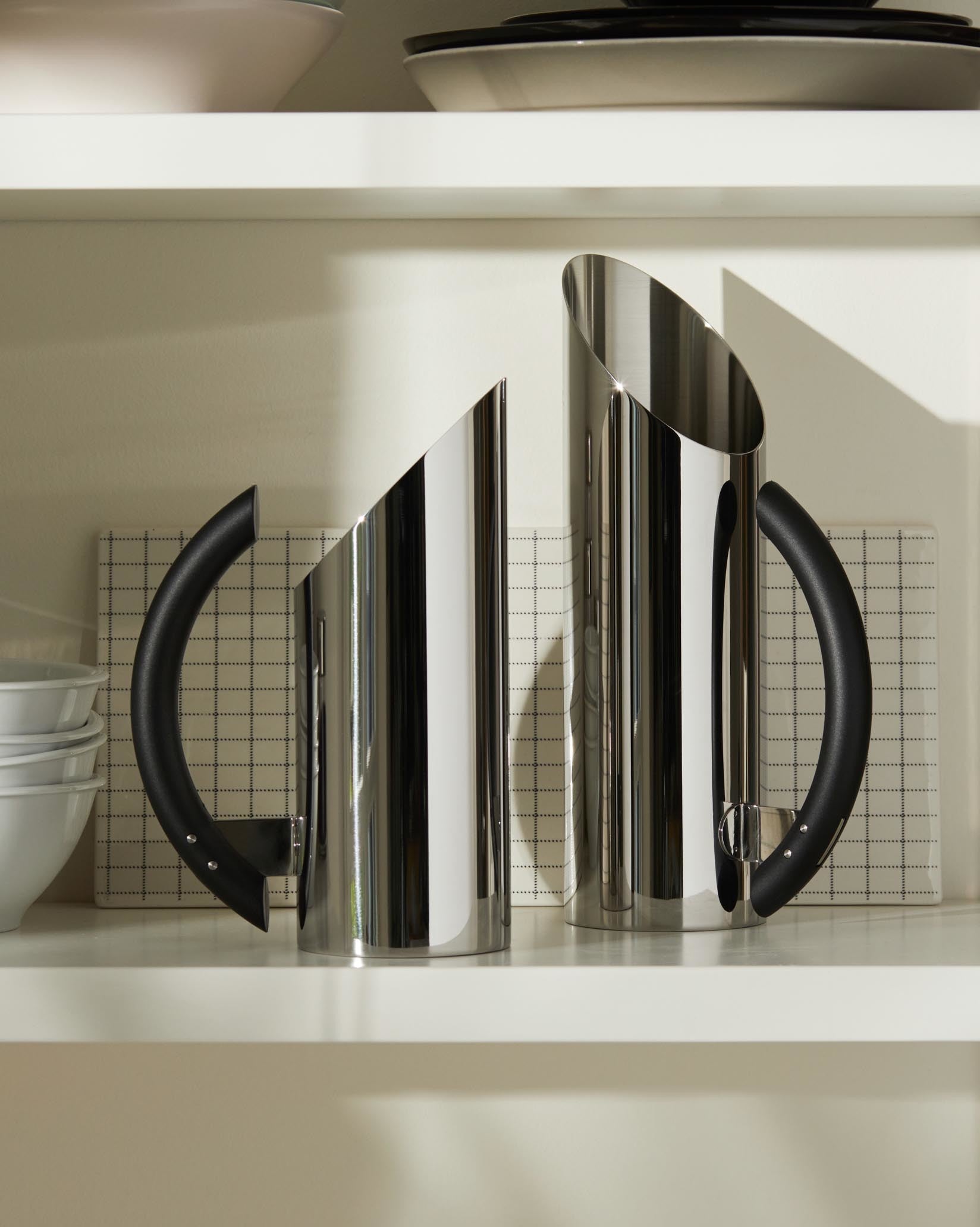 Two polished stainless steel pitchers by Alessi with angled tops and black handles, displayed on a white shelf.