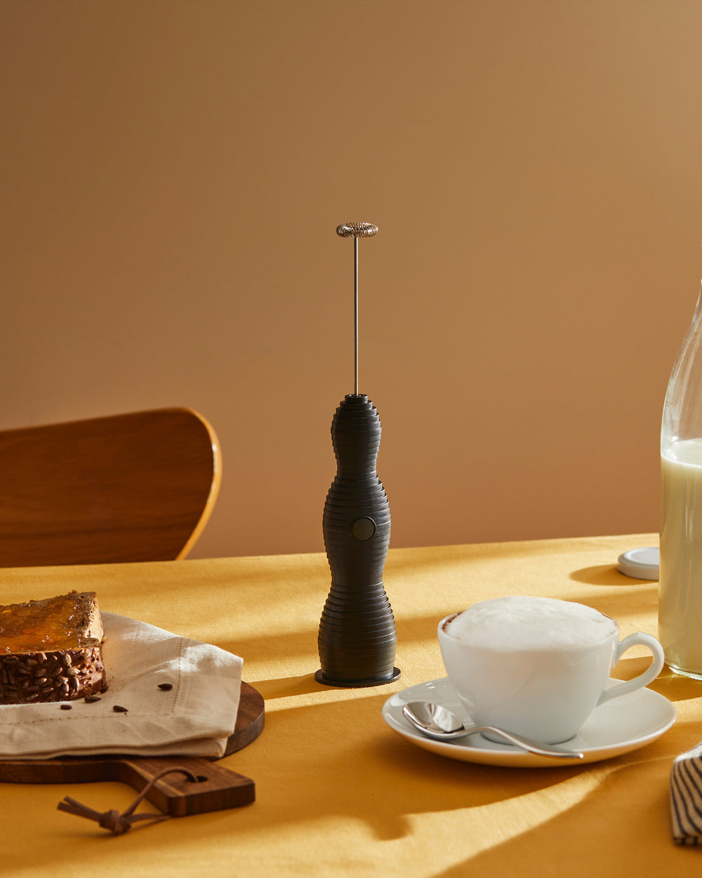 Black Pulcina handheld milk frother by Alessi with a ribbed design, standing on a yellow table beside a cup of frothed coffee and cake.