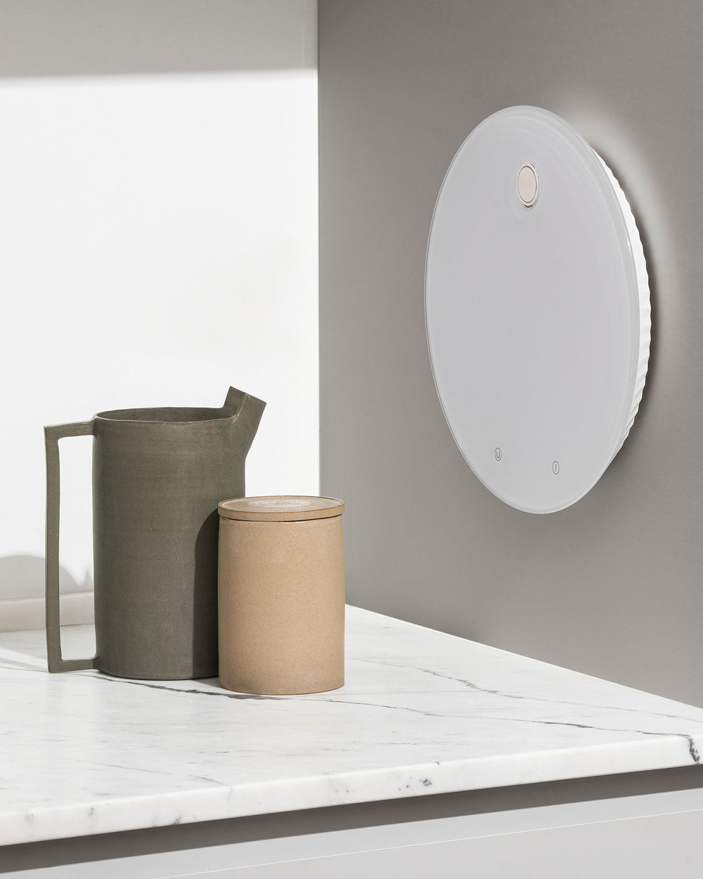 White circular electronic kitchen scales by Alessi with a sleek face and pleated sides, mounted on a gray kitchen wall.