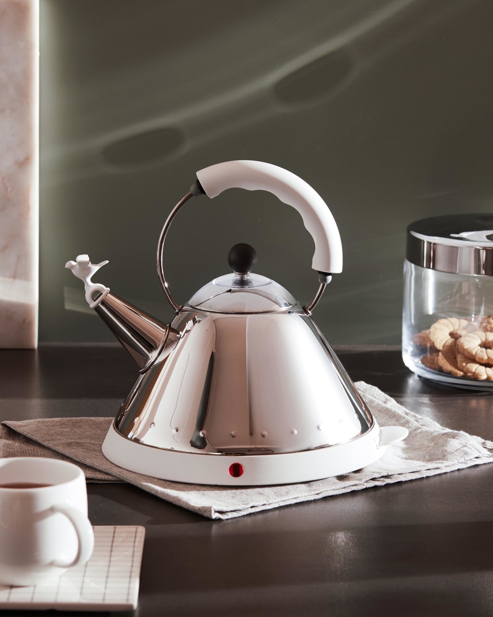 Silver conical kettle with white handle and base by Alessi, featuring a bird-shaped whistle on the spout and a red indicator light.