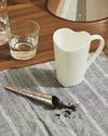 White heart-shaped cup by Alessi resting on a striped napkin, with a tea infuser and loose leaves.