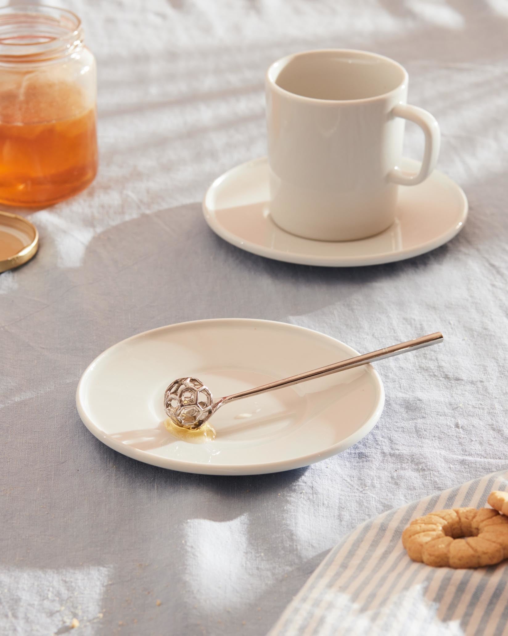 Acacia honey dipper, metallic with a spherical design, by Alessi, resting on a white plate with honey, a glass jar, white cup, and cookies.