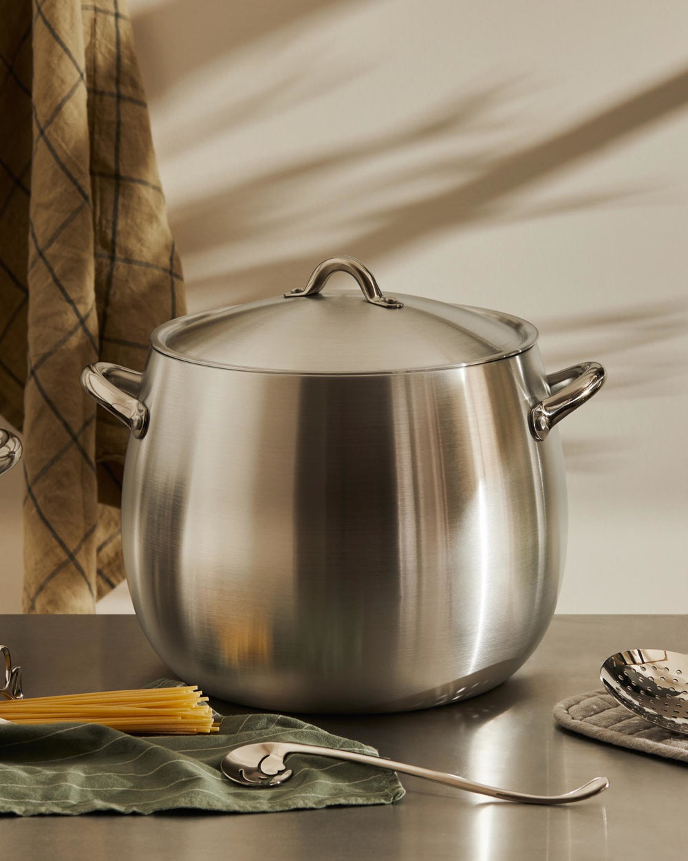 Large brushed stainless steel cooking pot by Alessi, with a lid and handles, on a metallic counter with spaghetti and a green striped cloth.