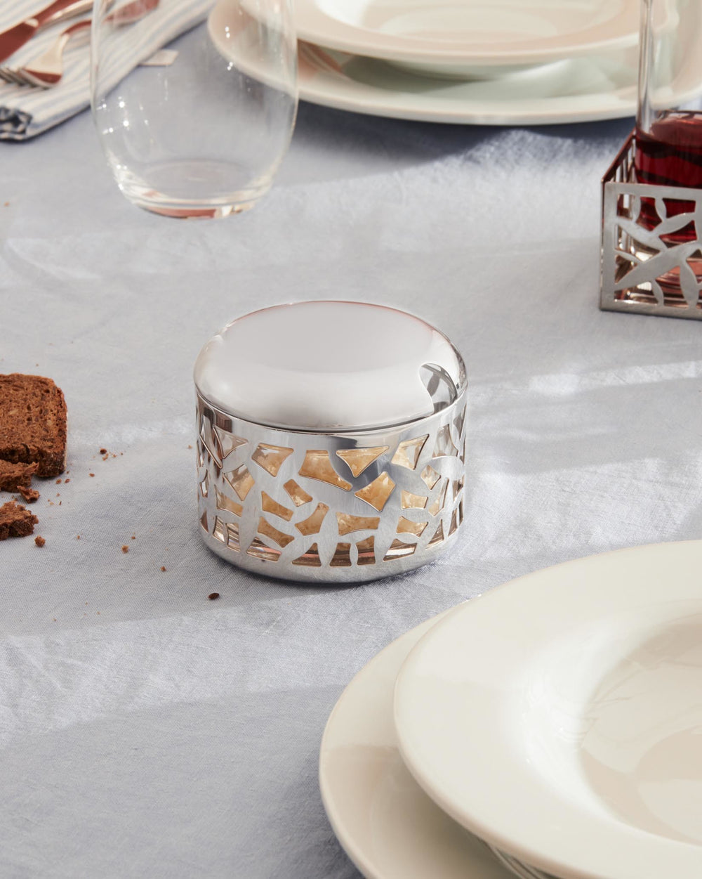 Silver-toned cheese cellar by Alessi with cactus-inspired cutouts, a domed lid, and spoon slot, on a light blue tablecloth.
