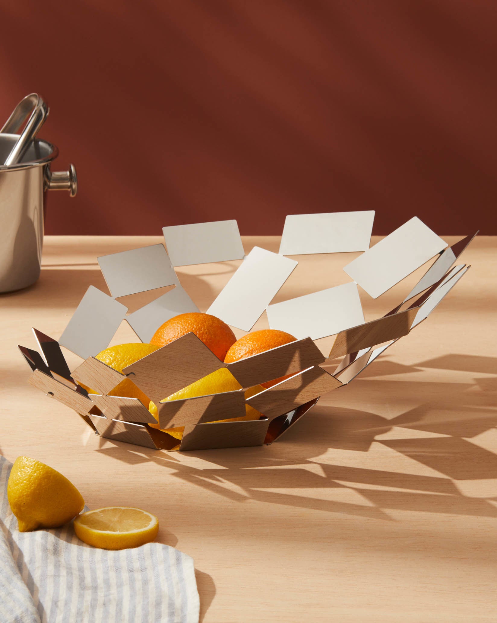 Geometric silver and wood fruit bowl by Alessi, holding lemons and oranges, on a light wood table.