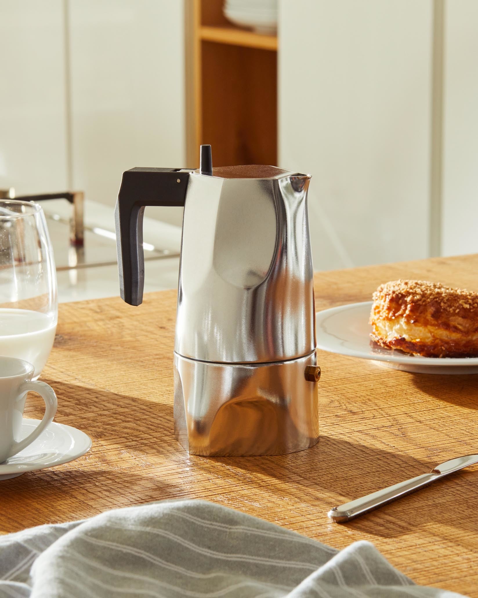Silver moka pot by Alessi with a black handle, on a wooden counter with breakfast items.