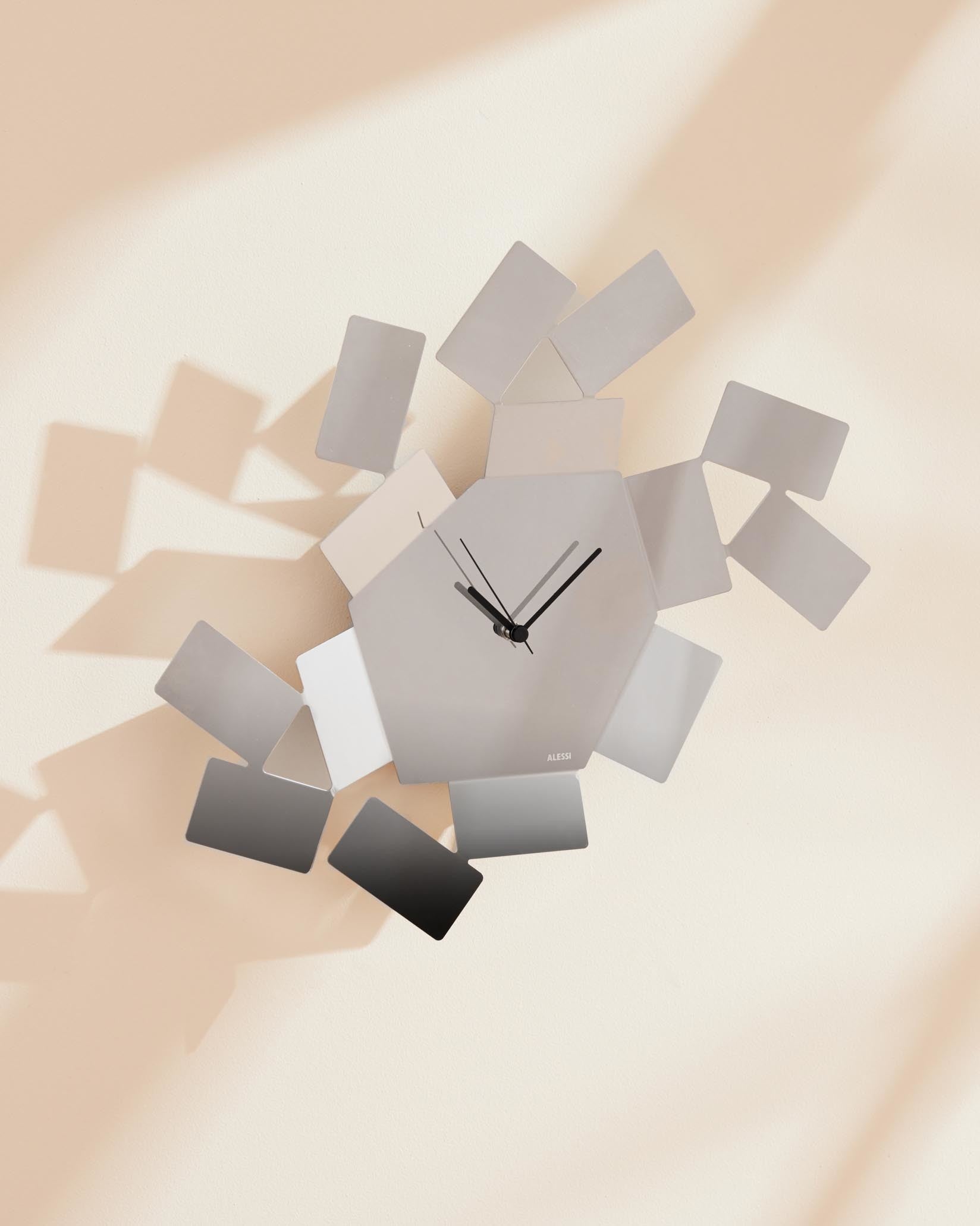 Modern hexagonal grey wall clock by Alessi, surrounded by scattered rectangular elements in varying shades of grey.