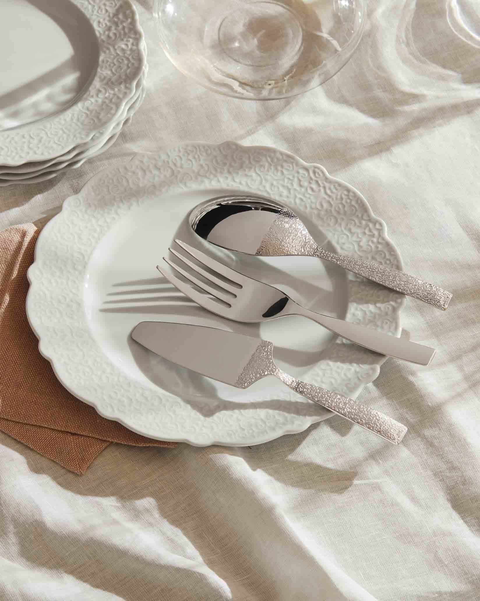 Silver patterned serving utensils by Alessi resting on an embossed white plate with a tan napkin and white tablecloth.