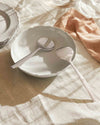 Silver serving cutlery set by Alessi, with a polished slotted spoon and a decorative textured spoon resting in a white bowl.
