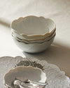 Scalloped white ceramic bowls by Alessi, stacked on a white tablecloth with decorative silver trays and shiny spoons.