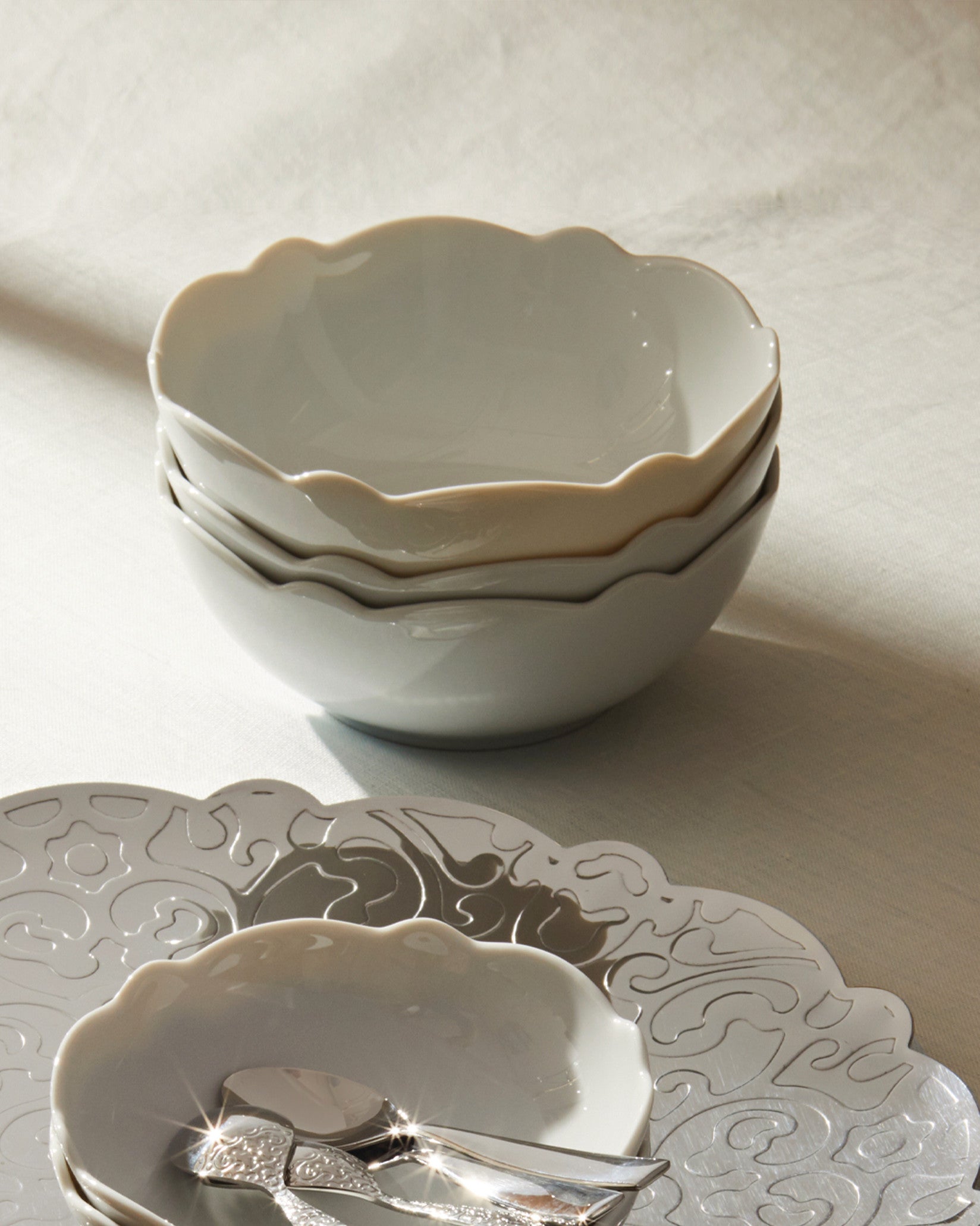 Scalloped white ceramic bowls by Alessi, stacked on a white tablecloth with decorative silver trays and shiny spoons.