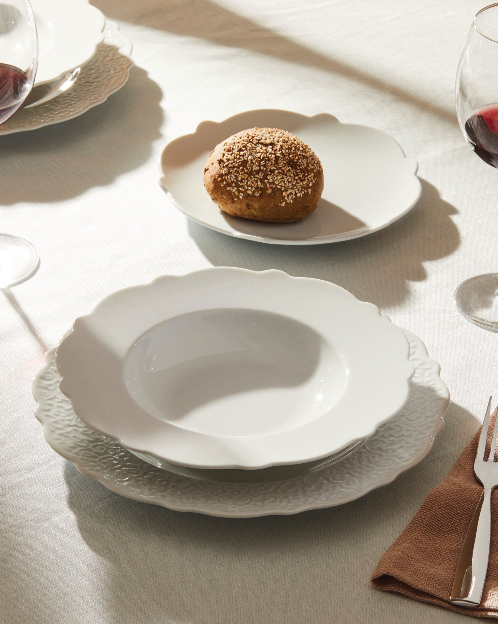 White deep plate with scalloped edge by Alessi, stacked on a patterned dinner plate with wine and bread.