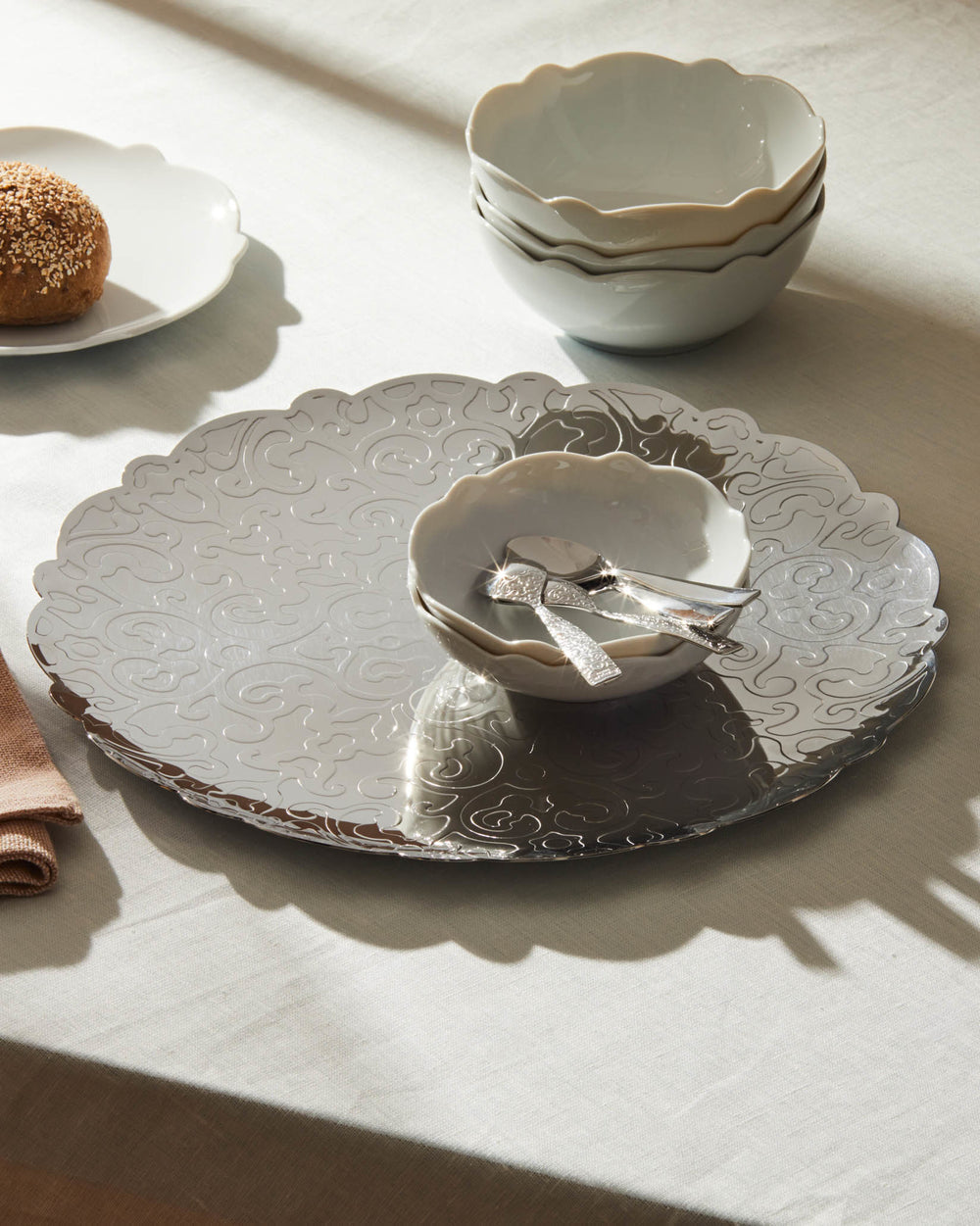 Silver scalloped serving platter by Alessi, with embossed scroll pattern, displayed with white bowls and ornate spoons.
