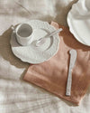 Embossed white porcelain and silver tableware by Alessi, including a coffee cup, saucer, spoon, and butter knife on a pink linen napkin.